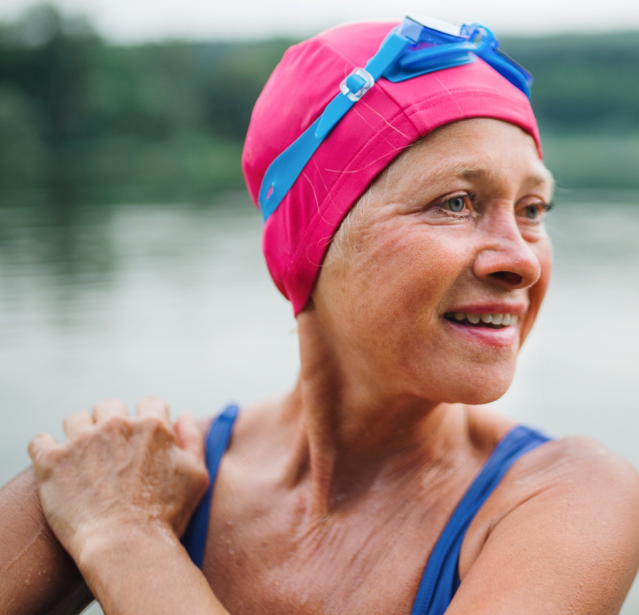 HCSC- A woman wearing a pink swim cap and blue swimsuit stands and smiles with a lake in the background